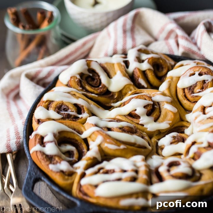 Freshly baked Gingerbread Cinnamon Rolls, generously drizzled with cream cheese icing, served in a rustic cast iron skillet with cinnamon sticks and golden forks in the background.
