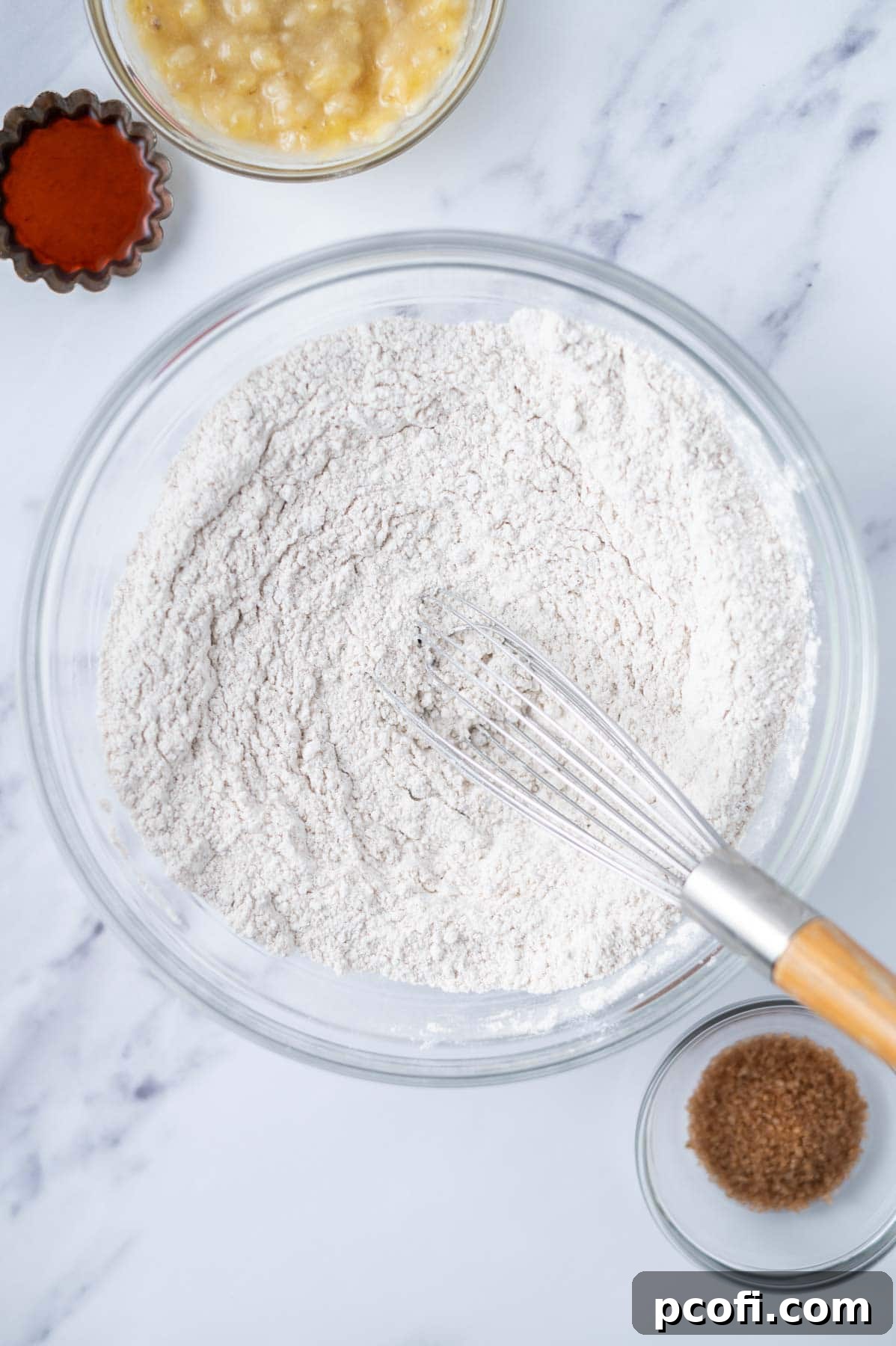Flour, baking powder, cinnamon, and salt are whisked together in a clear glass bowl with a metal whisk, ready for the next step in making banana scones.
