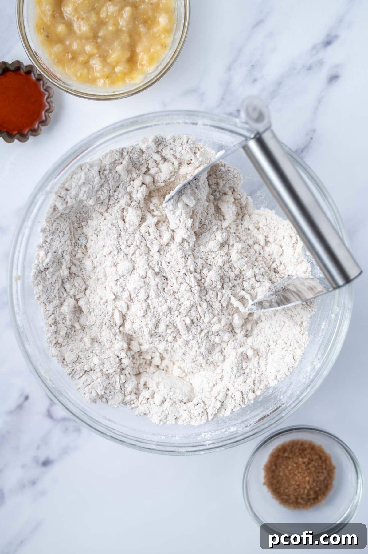 Grated frozen butter is being cut into a flour mixture in a glass bowl using a pastry cutter, creating a coarse meal texture for the scone dough.