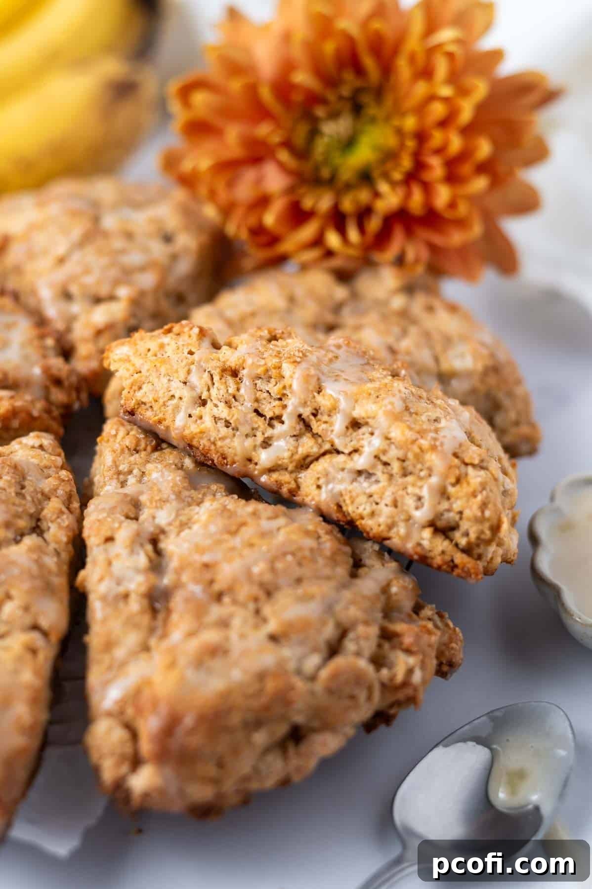 Beautifully glazed banana scones arranged on parchment paper, surrounded by fresh flowers and ripe bananas, highlighting their tempting appearance.