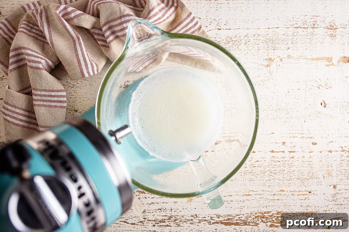 Granulated sugar and cornstarch being slowly streamed into whipping egg whites in a mixer bowl.