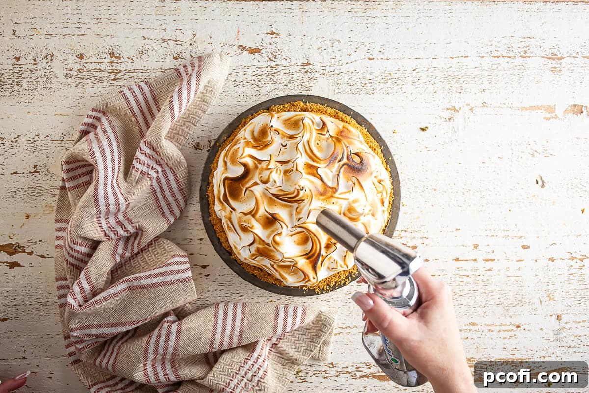 A kitchen torch being used to toast the meringue topping of an eggnog pie, creating golden-brown peaks.