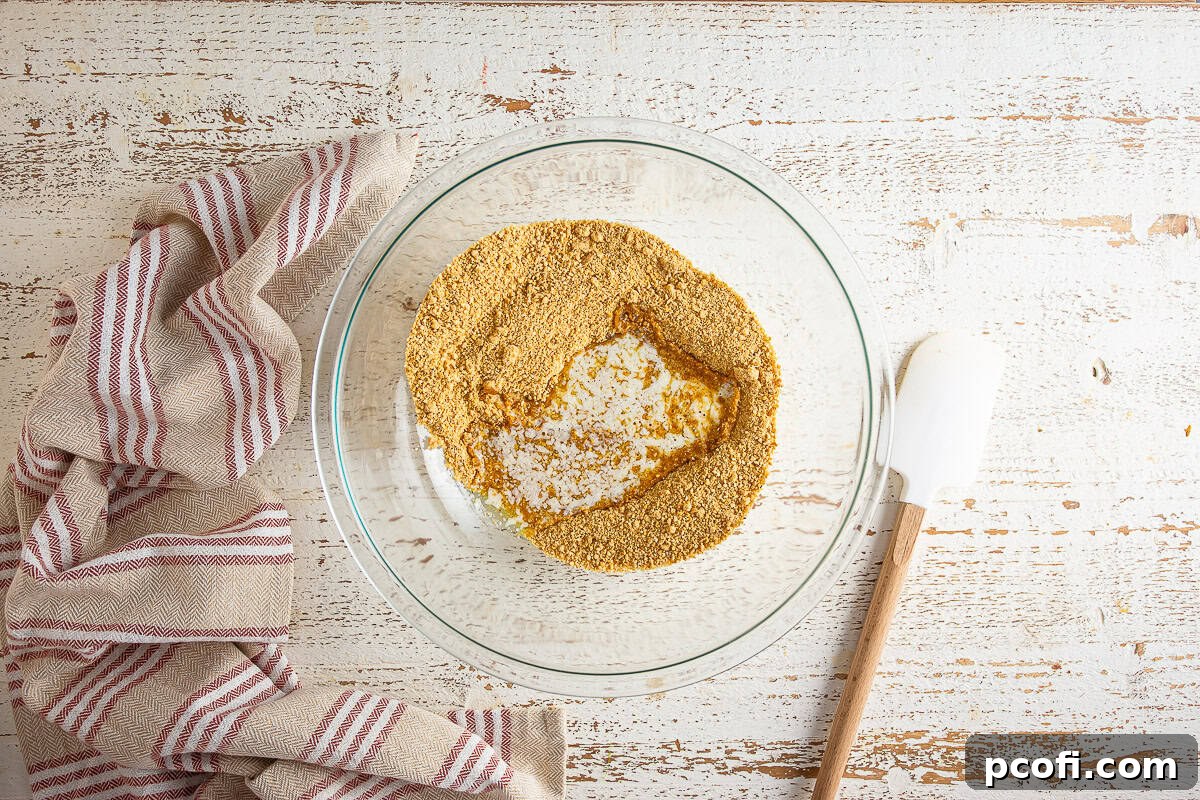 Graham cracker crumbs, brown sugar, cinnamon, and salt mixed in a glass bowl with a rubber scraper.