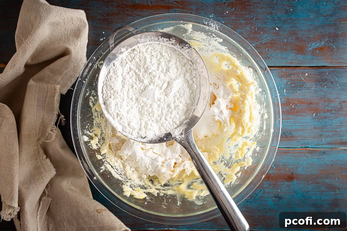A close-up shot of flour and cornstarch being sifted into the creamy alfajores dough mixture in a glass bowl, ensuring a smooth and lump-free dough.