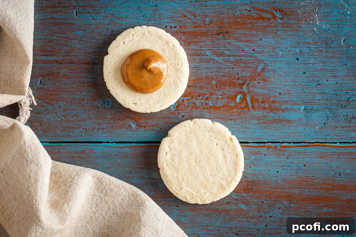 A delicate, baked alfajores cookie with a generous dollop of creamy dulce de leche perfectly centered on its flat surface, ready to be topped with another cookie.