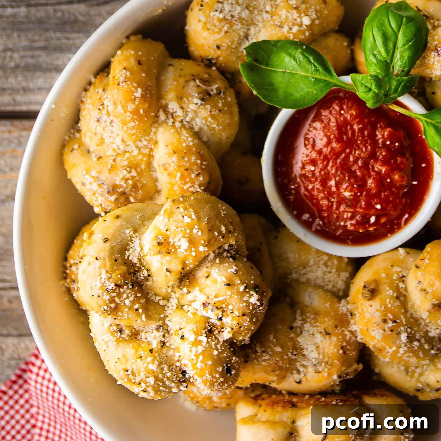 Golden-brown garlic knots in a bowl, garnished with fresh basil, and served with marinara sauce.