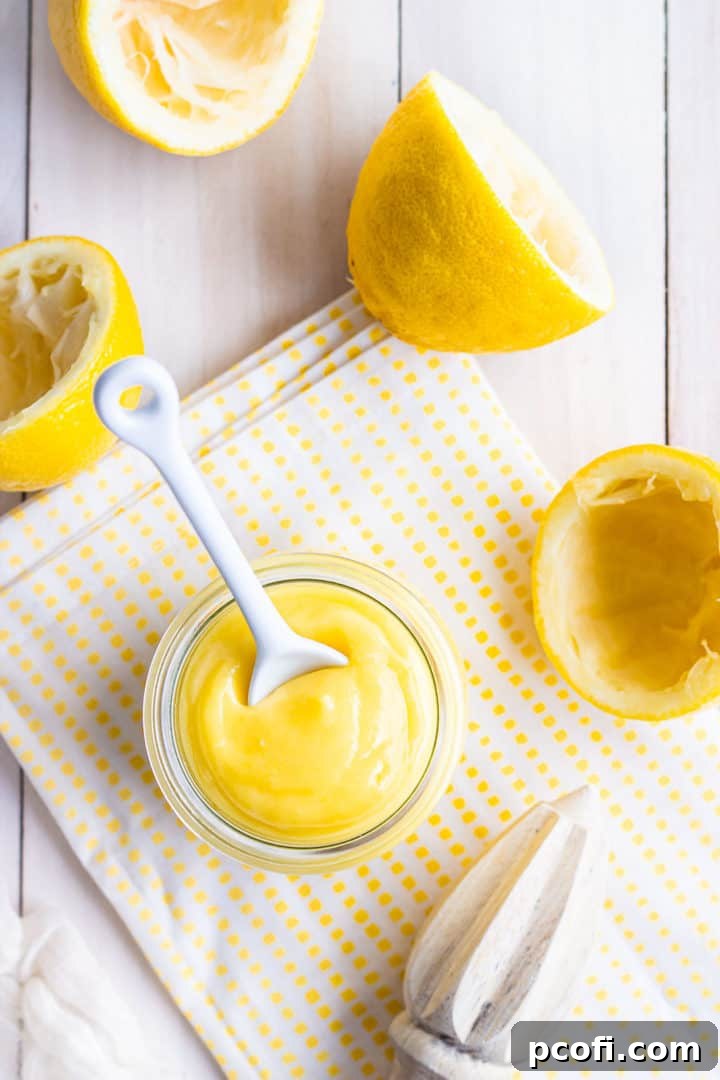 Overhead shot showcasing a clear glass jar of bright yellow lemon curd, surrounded by freshly cut lemons and a wooden citrus reamer on a vibrant yellow checkered cloth. This image perfectly captures the essence of fresh, homemade ingredients.