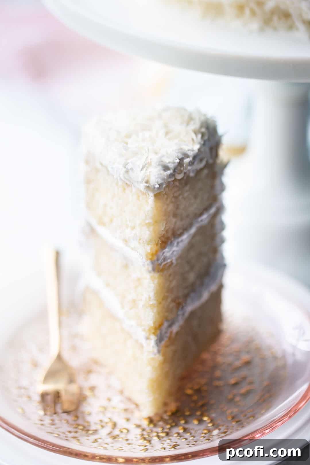 Tall wedge of coconut cream cake, served on a pink glass plate with a white pedestal in the background, showing off its delicate texture and layers.