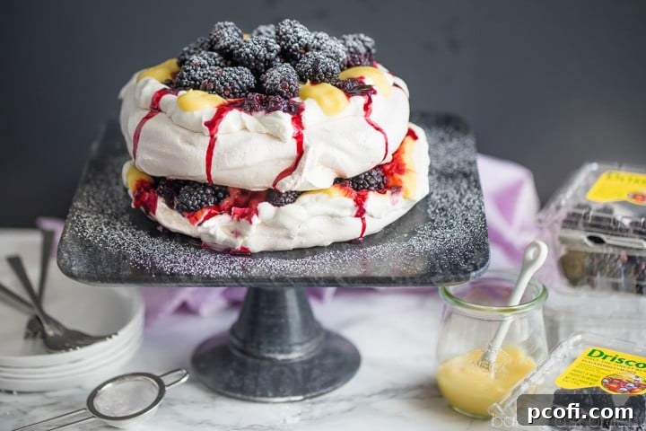A close-up side view of the Blackberry Lemon Pavlova, showcasing the distinct layers of crisp meringue, light whipped cream, glossy lemon curd, and fresh blackberries. The image emphasizes the dessert's impressive height and elegant presentation, highlighting the use of Driscoll's #FinestBerries.