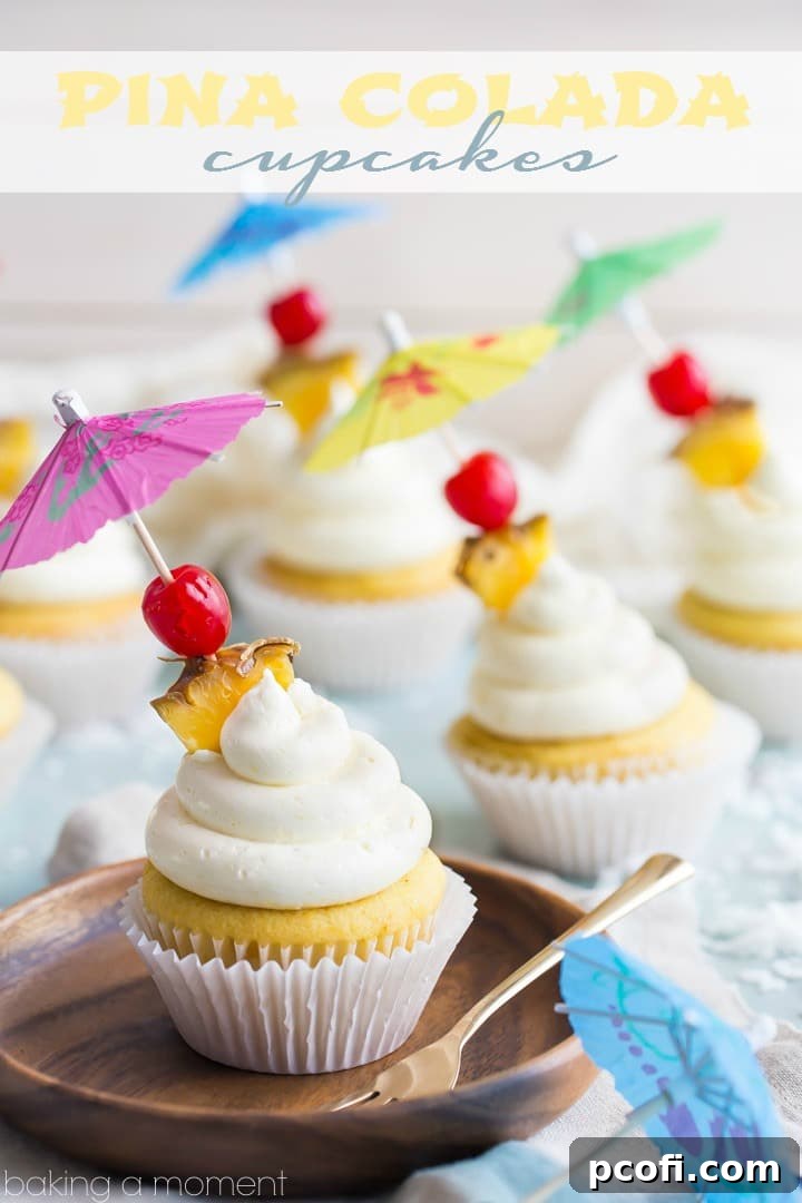Close-up of a perfectly frosted Pina Colada cupcake, showcasing the creamy white buttercream, a bright red maraschino cherry, and a small pineapple chunk, against a blurred background of more cupcakes.