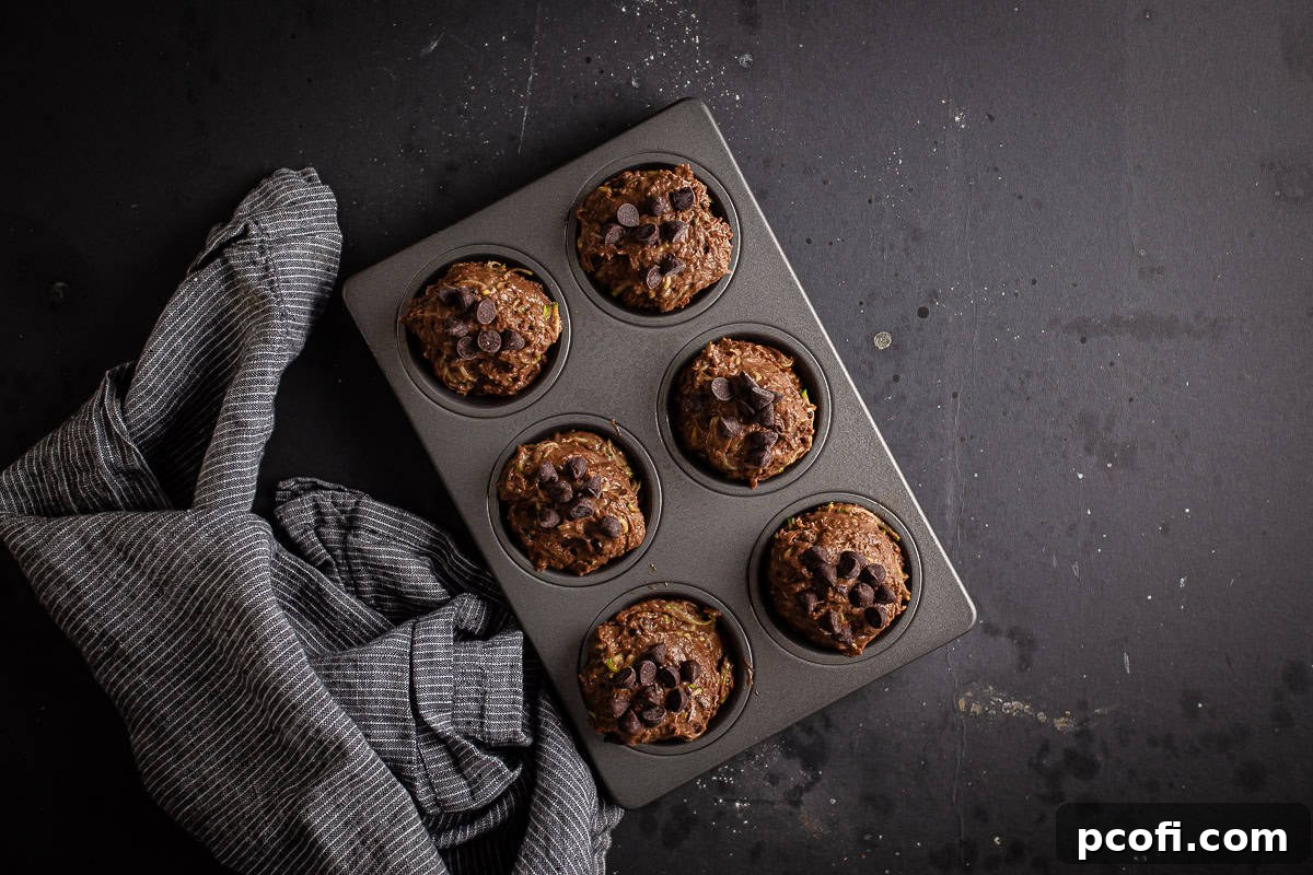 Unbaked chocolate zucchini muffins in a tin with a striped kitchen towel.