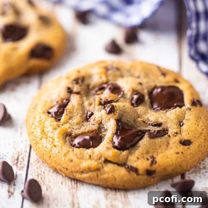 Close up image of a chocolate chip cookie on a distressed white table, surrounded by chocolate chips, showcasing its soft texture and gooey chocolate.