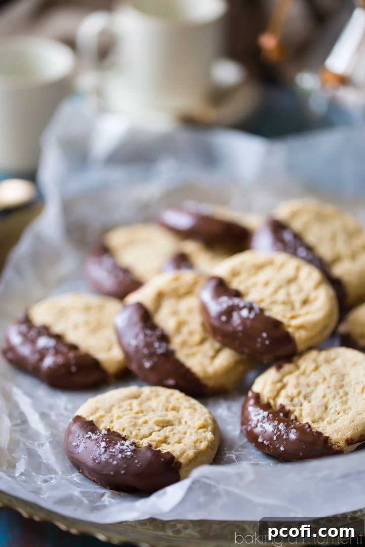 A hand gently holding a freshly baked, soft almond butter cookie. One side is generously coated in glossy dark chocolate and sprinkled with vibrant sea salt flakes, ready to be enjoyed as a perfect sweet and salty treat.