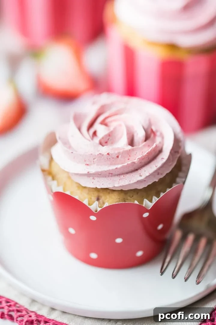 A trio of beautifully decorated strawberry cupcakes with strawberry buttercream, presented in charming pink, red, and white cupcake wrappers. They sit atop a pristine white cake stand, with a subtle pink-patterned cloth in the background, exuding elegance and deliciousness.