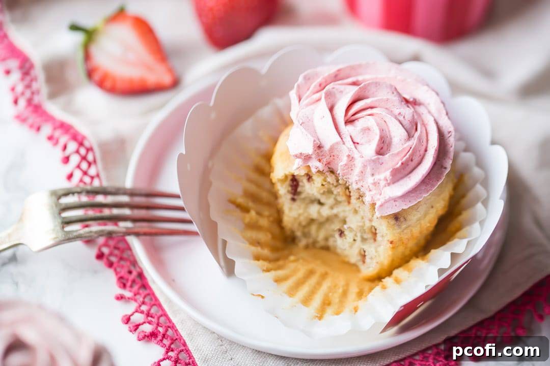An array of exquisite strawberry cupcakes, generously topped with fluffy strawberry buttercream, neatly arranged in decorative pink, red, and white liners on a classic white cake stand. The background features a delicately patterned pink cloth, enhancing the visual appeal of these fresh and flavorful treats.