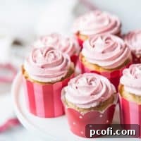 Strawberry cupcakes with strawberry buttercream, in pink, red, and white cupcake papers, sitting on a white cake stand. The buttercream has been piped in the shape of a rosette, and there is a pink-trimmed cloth in the background.
