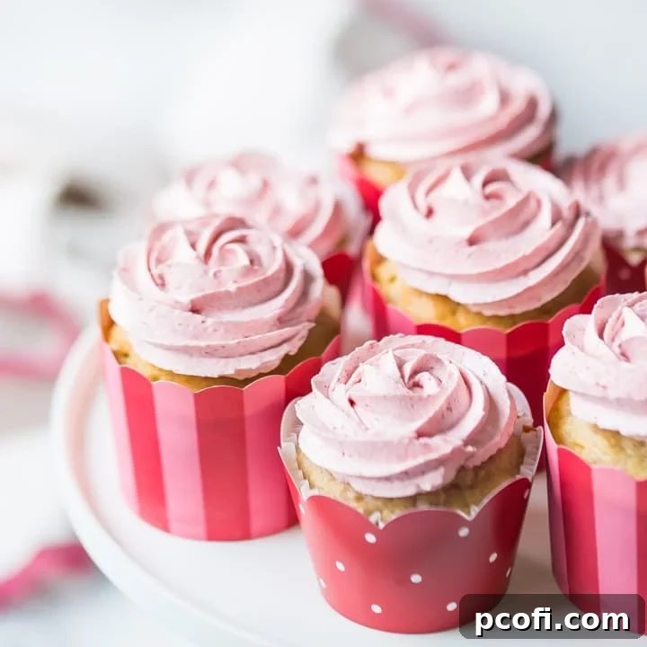 Luscious strawberry cupcakes topped with strawberry Swiss meringue buttercream, arranged on a white cake stand. The buttercream is expertly piped into beautiful rosettes, with a soft pink-trimmed cloth in the background, highlighting their fresh, natural appeal.