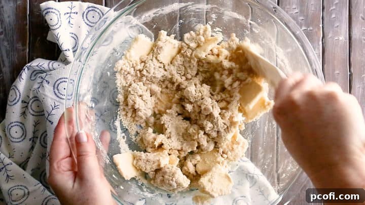 Stirring crumbly pie dough in a large glass bowl with a silicone spatula, checking for proper consistency before chilling.