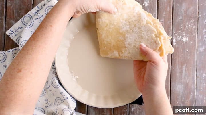 Carefully transferring folded pie crust dough into a pie dish, ensuring not to stretch it.