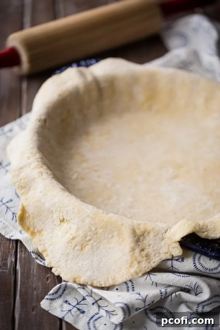 Vertical image of fresh homemade pie dough gently draped over a pie pan, with a rolling pin and flour in the background.