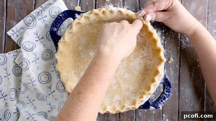 Close-up of hands crimping the edge of an unbaked pie crust in a dish, creating a decorative border.
