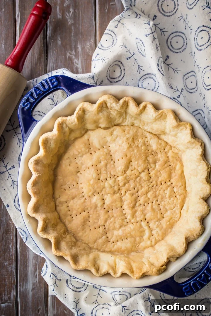 Vertical overhead image of a golden-brown, flaky baked pie crust in a dark blue dish, with a rolling pin nearby.