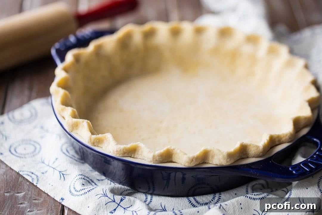 Horizontal image of an unbaked pie crust in a dark blue dish, with a rolling pin resting nearby on a floured surface.