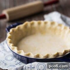 Square image of unbaked pie crust in a dark blue dish with a rolling pin in the background.