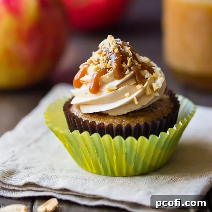 Delicious caramel apple cupcakes on a rustic wooden stand, showcasing caramel buttercream, a caramel drizzle, and chopped peanuts. Fresh apples and whole peanuts are in the soft-focus background, enhancing the autumn theme. Keywords: food, desserts, apple, fall baking.