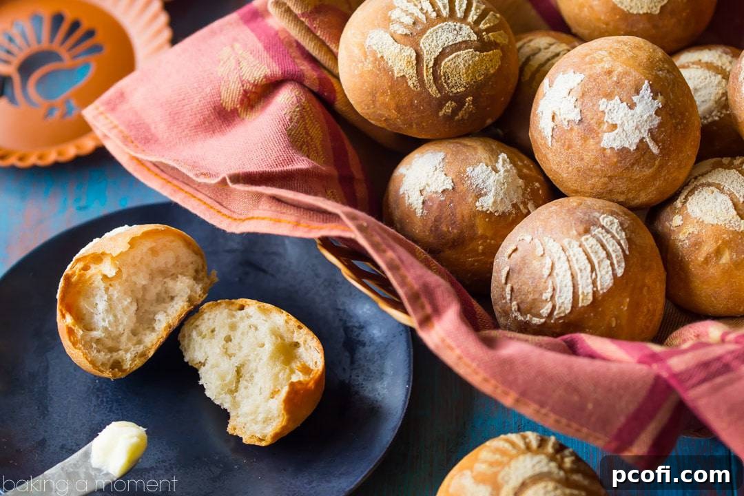A crusty bread roll, split in half, on a plate with a butter knife and a basket of fall-themed stenciled bread rolls in the background.