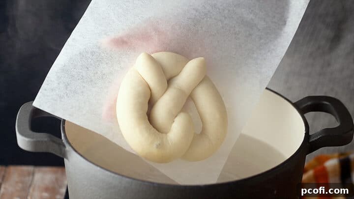 A homemade soft pretzel, still on its parchment paper, gently being lowered into a simmering baking soda bath.