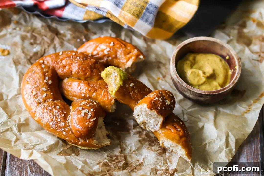 A perfectly crusty, golden-brown homemade soft pretzel, torn in half and ready for dipping, with one end immersed in spicy brown mustard, all resting on crumpled parchment paper.