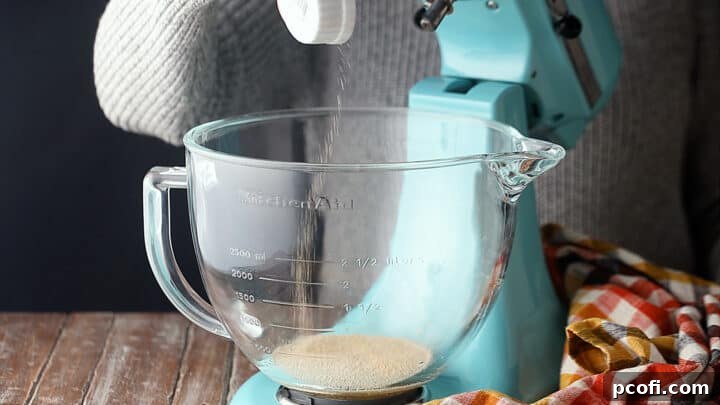 Dry active yeast being sprinkled over warm water and brown sugar in a clear glass bowl, preparing for activation.