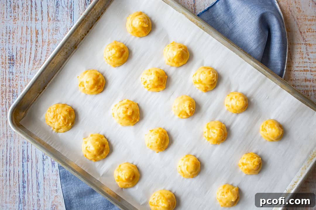 Unbaked cheese puffs, precisely piped onto a parchment-lined baking sheet, ready for the oven.