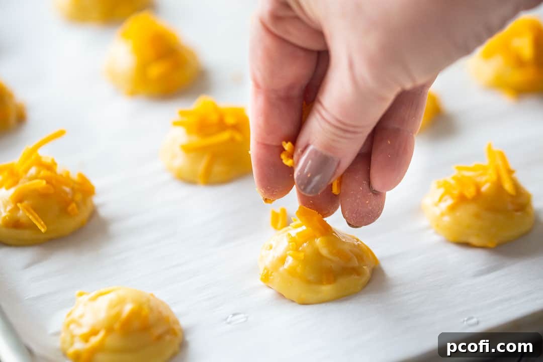 A hand sprinkling extra cheese over unbaked choux puffs on a baking sheet, preparing them for the oven.