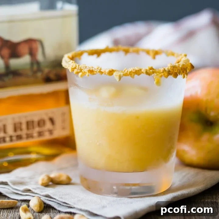 Caramel apple slush cocktail in a rocks glass with caramel and crushed peanuts around the rim. Fresh apple and a bottle of bourbon in the background, with peanuts scattered in the foreground. 