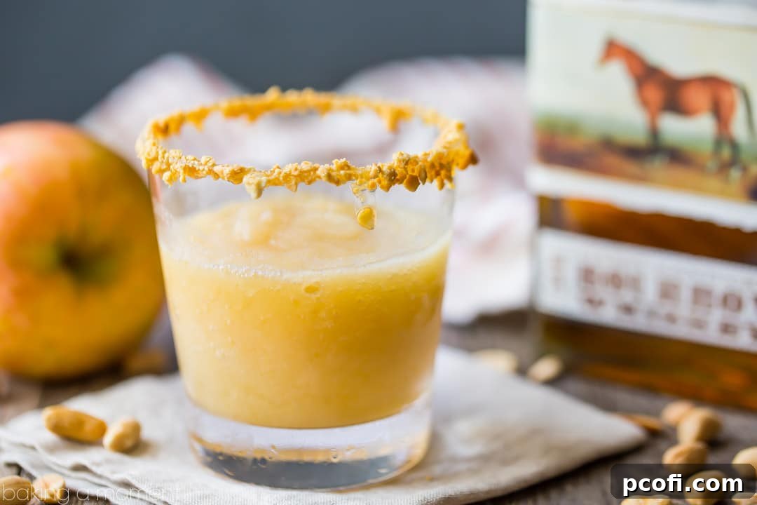 Caramel apple bourbon slush in a rocks glass with a caramel & crushed peanut crusted rim. Bourbon bottle in the background with fresh apple and peanuts scattered on the table. 