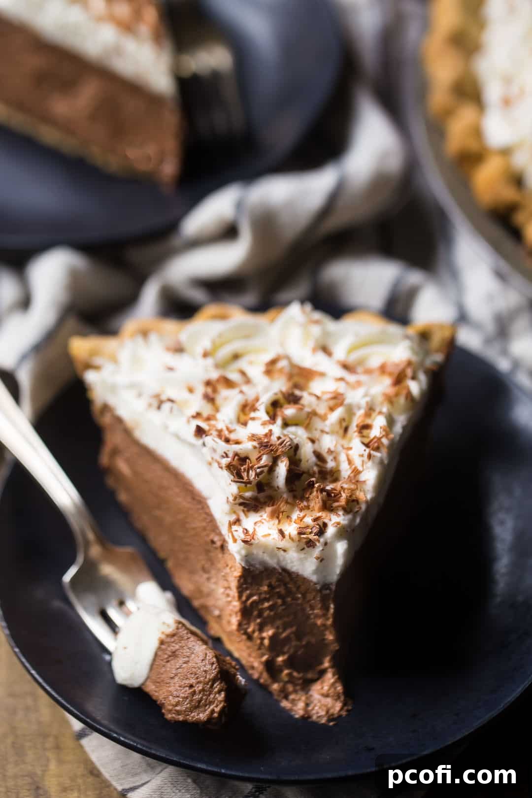 A generously thick slice of homemade chocolate cream pie, topped with a luscious swirl of whipped cream and delicate chocolate shavings, is presented against a dark background, showcasing its rich layers.