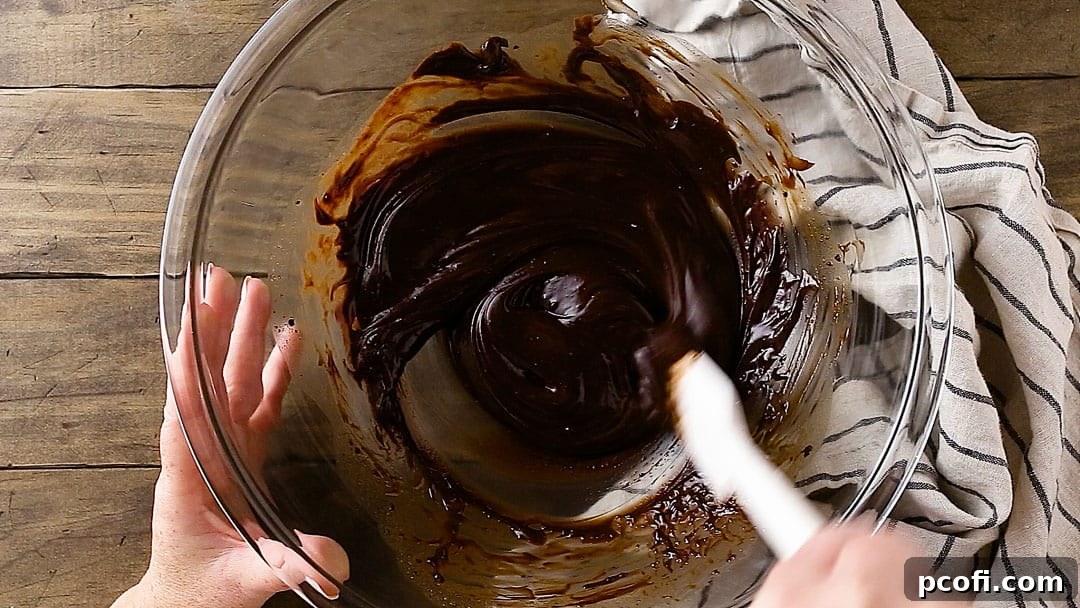 A large mixing bowl containing the melted chocolate mixture is being gently stirred with a spatula, creating a smooth and glossy texture, indicating it's ready for the next step.