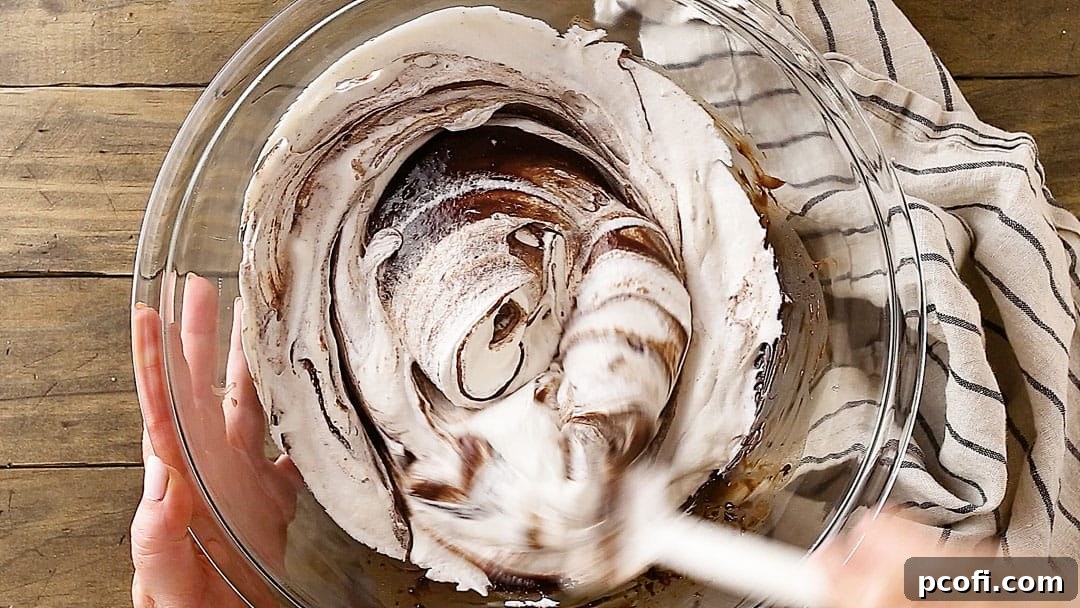 Melted chocolate is being delicately folded into a bowl of whipped cream, ensuring an even, airy blend for the light and fluffy chocolate pie filling.