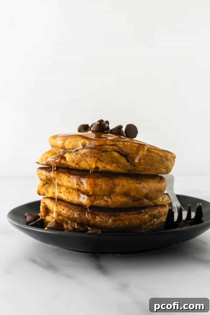 A beautifully stacked plate of golden pumpkin pancakes, ready for a delicious breakfast, with a fork resting beside them on a sleek black plate.