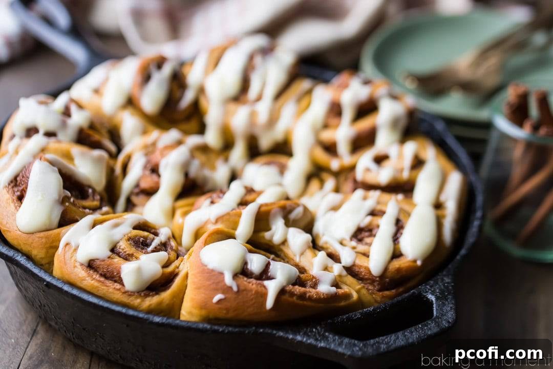 Iron skillet filled with gingerbread cinnamon rolls drizzled with cream cheese icing. Cinnamon sticks and stack of green plates in the background.