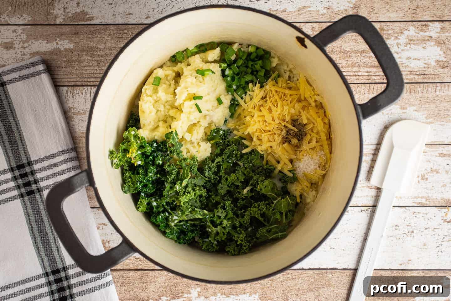 Various ingredients, including chopped kale, scallions, shredded cheese, and hot milk, being added to a pot of mashed potatoes.