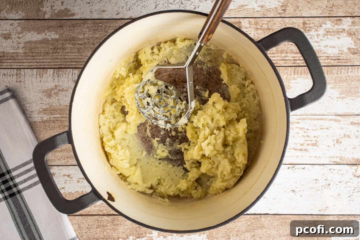 A potato masher actively mashing boiled potatoes and butter in a large pot, creating a creamy base.