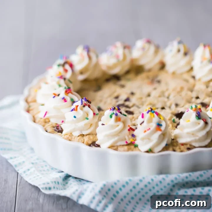 A beautifully decorated giant chocolate chip cookie cake with vanilla buttercream rosettes and vibrant rainbow sprinkles, set on a turquoise cloth with a gray background.