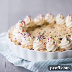 Square image of a giant chocolate chip cookie cake with vanilla buttercream rosettes and rainbow sprinkles, with a turquoise cloth on a gray background.
