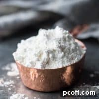 Square image of a copper measuring cup filled with cake flour, on a dark background.