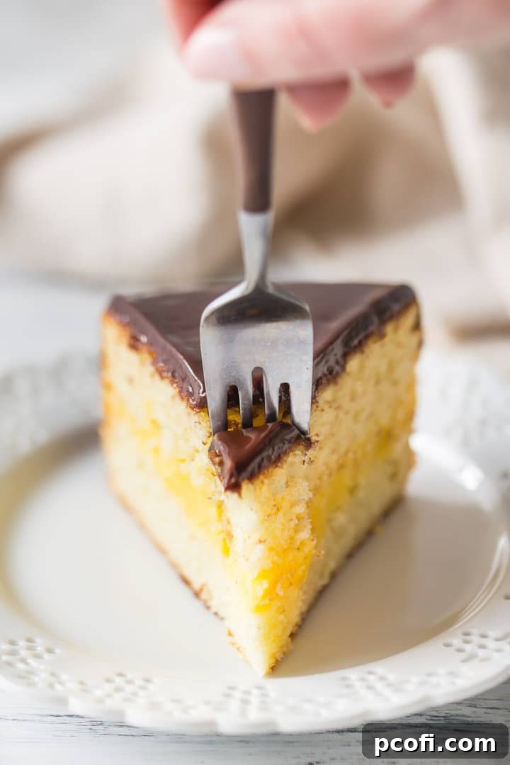 A fork gently sinks into a slice of homemade Boston Cream Pie, revealing the fluffy vanilla cake and creamy filling layers beneath the shiny chocolate glaze, ready to be enjoyed.