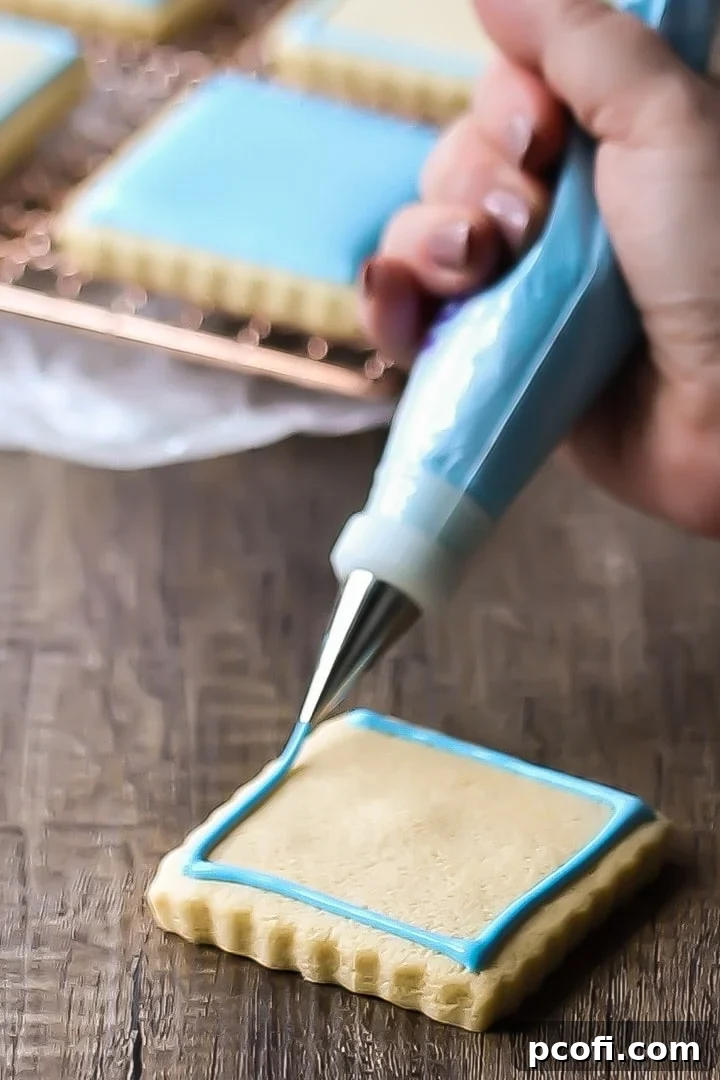 Close-up of a pastry chef meticulously piping a thin, precise line of blue royal icing along the border of a square sugar cookie, preparing it for flooding.