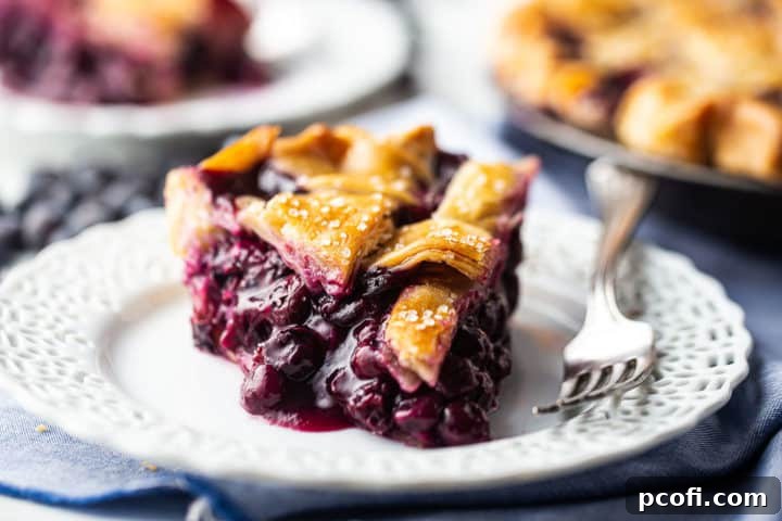 Close-up image of blueberry pie filling recipe, baked into a flaky pie crust and served on a white plate with a blue napkin.